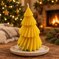 A yellow tiered tree-shaped candle sits on a small beige plate on a wooden table, with a cozy holiday scene in the background including a lit fireplace, Christmas tree lights, and pine branches.