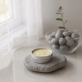 Silver tin with bee balm with beeswax, cocoa and shea butter on a  gray stone coaster on a white surface with a plant in the background.