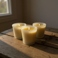 Three beeswax votive candles in glass holders on a wooden surface with natural light.