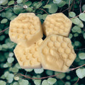 Three hexagonal honeycomb-shaped solid lotion bars on a green leafy background