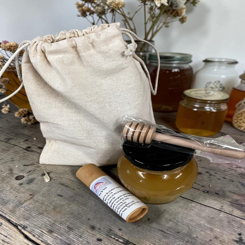 Honey jar with dipper, drawstring bag, and honey sticks on a wooden surface.